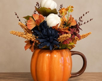 May include: A ceramic pumpkin-shaped mug in bright orange with a brown handle, filled with an arrangement of artificial flowers including dark blue dahlias, orange blossoms, yellow wheat-like stems, and two small white decorative pumpkins.