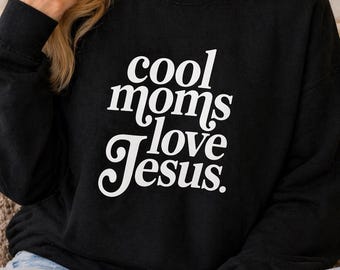 Puede incluir: Sudadera negra con el texto blanco "cool moms love Jesus." El texto está en una fuente elegante y cursiva. La sudadera es de cuello redondo y parece estar hecha de un material suave.