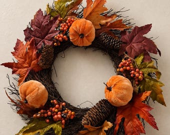Puede incluir: Una corona circular hecha de ramas de color marrón oscuro, decorada con tres calabazas de terciopelo naranja, racimos de pequeñas bayas naranjas y rojas, piñas marrones y un surtido de hojas de arce de colores otoñales en tonos rojo, naranja y verde.