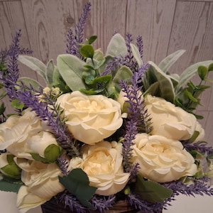 May include: A floral arrangement featuring cream-colored roses, purple lavender, and green foliage. The flowers are arranged in a brown woven basket with a burlap bow. The background is a light wood-paneled wall.