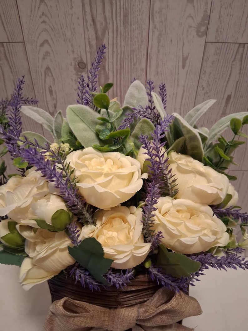 May include: A floral arrangement featuring cream-colored roses, purple lavender, and green foliage. The flowers are arranged in a brown woven basket with a burlap bow. The background is a light wood-paneled wall.