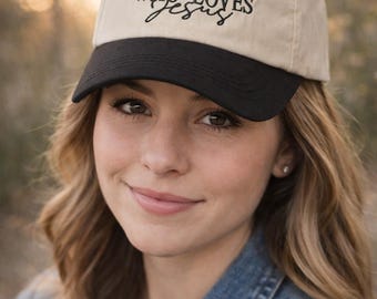 Puede incluir: Gorra de béisbol beige y negra con el texto "JUST A GIRL WHO LOVES Jesus" en escritura negra. La gorra tiene una visera curva y un botón negro en la parte superior. La persona de la imagen lleva una chaqueta vaquera.