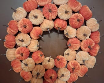 Puede incluir: Una corona circular compuesta por numerosas calabazas pequeñas cubiertas de tela en varios tonos de naranja y beige. Cada calabaza presenta un tallo marrón oscuro y una superficie texturizada suave, dispuestas densamente en un anillo.