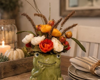 May include: A glossy green ceramic frog planter featuring a smiling face and a hand raised to its chin. The planter is filled with an arrangement of artificial flowers in shades of orange, red, and white, accented with brown stalks and green leaves.
