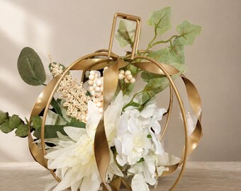 May include: A decorative pumpkin-shaped sculpture crafted from curved gold-toned metal ribbons. The hollow center is filled with an arrangement of artificial white flowers, green eucalyptus leaves, small cream-colored berries, and light green ivy.