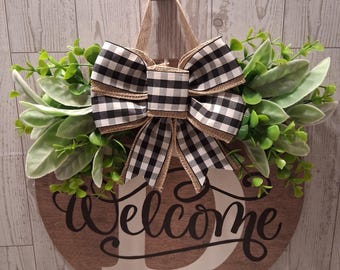 Puede incluir: Un letrero de puerta redondo de madera con acabado de veta de madera marrón que presenta la palabra Welcome en letra cursiva negra sobre una letra D blanca grande. El letrero está decorado con un lazo a cuadros blanco y negro y follaje verde.