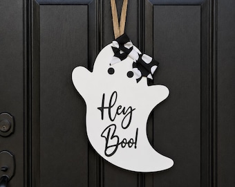 Puede incluir: Un letrero de puerta con forma de fantasma blanco que presenta el texto negro "Hey Boo!" en letra cursiva. El fantasma tiene dos ojos circulares negros y está adornado con un lazo de lunares blancos y negros, colgado de un lazo de arpillera.