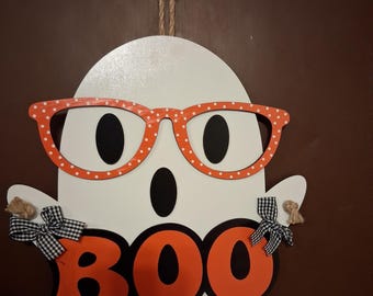 Puede incluir: Un adorno de pared con forma de fantasma blanco que presenta gafas naranjas con lunares blancos, ojos ovalados negros y una boca negra. La palabra BOO está escrita en letras naranjas grandes con bordes negros. Incluye dos lazos de cuadros vichy.