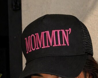 Puede incluir: Gorra de camionero negra con la palabra "MOMMIN'" bordada en rosa en la parte delantera. La gorra tiene una visera curva y una parte trasera de malla. Un accesorio de moda.