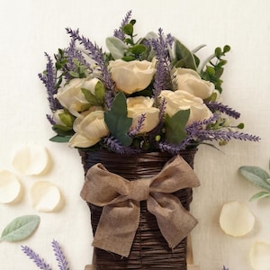 May include: A floral arrangement featuring cream roses, soft lavender, and greenery in a woven basket with a burlap bow. The text reads: "A touch of handcrafted elegance, a beautiful arrangement of cream roses, soft lavender and rustic greenery."
