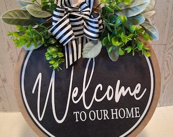 Puede incluir: Cartel de madera redondo con las palabras "Welcome TO OUR HOME" en escritura blanca sobre un fondo negro. El cartel está decorado con follaje artificial verde y gris y un lazo a rayas blanco y negro. Se muestra sobre un pequeño caballete de madera.