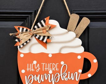 Puede incluir: Un colgador de puerta con forma de taza naranja con lunares blancos y el texto "HEY THERE pumpkin" en letra blanca. Incluye una cobertura de nata montada blanca, dos ramas de canela de madera y un lazo decorativo de tela a rayas.
