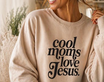 Puede incluir: Una sudadera beige con el texto negro "cool moms love Jesus". La sudadera tiene cuello redondo y mangas largas. La imagen muestra a una persona que lleva la sudadera.