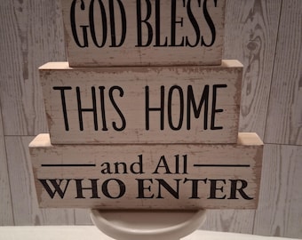 Puede incluir: Un letrero de bloques de madera apilados con las frases "GOD BLESS", "THIS HOME" y "and All WHO ENTER" en letras negras. Los bloques son de color blanquecino y están sobre un pedestal blanco.