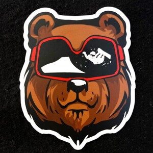 May include: A sticker featuring a brown bear wearing red-framed ski goggles. The goggles reflect a snowy mountain scene with a skier. The bear has a white outline and a detailed design, perfect for decorating laptops, water bottles, or other items.