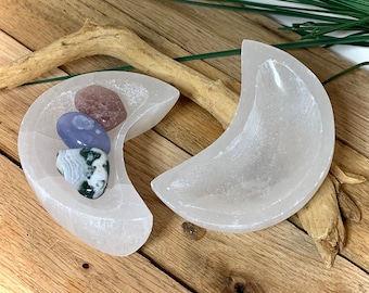 May include: Two crescent moon-shaped bowls carved from translucent white selenite. One bowl contains three smooth, polished stones: one pink, one blue, and one green and white patterned stone. The bowls have a smooth, matte finish.