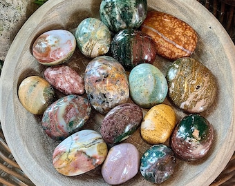 May include: A collection of smooth, polished, multi-colored stones resting in a shallow, light-colored wooden bowl. The stones feature various patterns and hues, including shades of green, brown, red, yellow, and purple, with glossy surfaces.