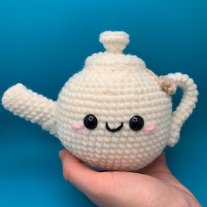 May include: A white crocheted teapot with a smiling face and black eyes. The teapot has a small handle and a spout.