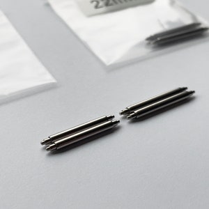 May include: A close-up of six silver watch band spring bars. The spring bars are arranged in two groups of three, with the ends of the bars facing each other.