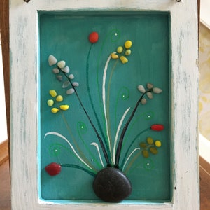 Puede incluir: Una obra de arte enmarcada que presenta un fondo turquesa pintado con una piedra negra y flores de guijarros de colores. Las flores están hechas de pequeñas piedras de colores y están dispuestas en forma de jarrón. La obra de arte está enmarcada en madera blanca.
