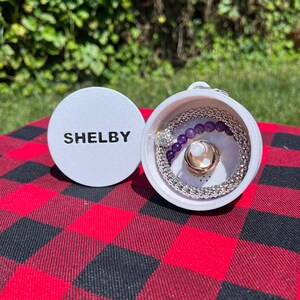 Op de afbeelding: Een witte ronde container met het woord "SHELBY" gedrukt op de deksel. De container is open en bevat een zilveren kralenarmband, een paarse kralenarmband en een gouden ring.