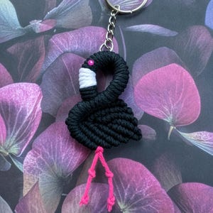 Puede incluir: Llavero de flamenco de macramé negro con un llavero plateado. El flamenco tiene un pico blanco, un ojo rosa y patas rosas. El fondo es un estampado floral morado oscuro.