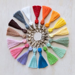 May include: A set of 18 colorful tassel keychains with geometric wooden beads. Each keychain features a different color tassel and a matching wooden bead.