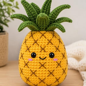 Może przedstawiać: Żółta szydełkowana pluszowa zabawka w kształcie ananasa z zielonymi liśćmi na górze. Ananas ma uśmiechniętą buzię z dwojgiem okrągłych czarnych oczu i małymi różowymi wyszywanymi policzkami. Ciało ma teksturowany wzór krzyżykowy z brązowej włóczki.