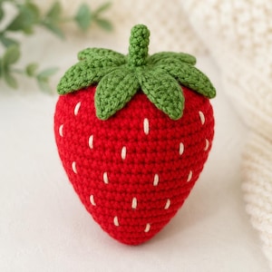 May include: A handcrafted crochet strawberry toy featuring a vibrant red body with small white stitched details representing seeds. The top of the strawberry is adorned with a textured green crocheted stem and a cluster of green leaves.
