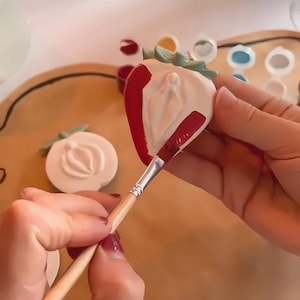 May include: A person is painting a ceramic strawberry with red paint using a wooden paintbrush. The strawberry has a white glaze and a green stem. The strawberry is sitting on a brown surface.