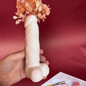 Puede incluir: Un jarrón de cerámica rosa claro con forma de pene, que contiene flores secas de color melocotón. El jarrón tiene texturas superficiales detalladas. Un juego de pintura con pinceles y una paleta de colores está en primer plano.