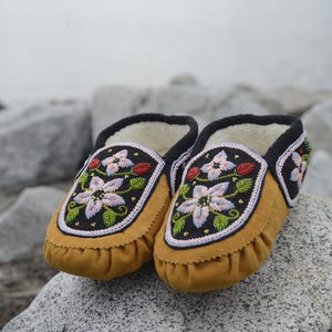 May include: A pair of brown leather moccasins with floral embroidery. The moccasins have a black trim and are decorated with pink, white, and green flowers.