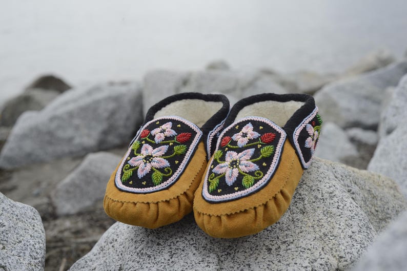 May include: A pair of brown leather moccasins with floral embroidery. The moccasins have a black trim and are decorated with pink, white, and green flowers.