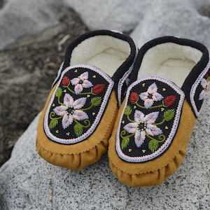 May include: A pair of brown leather moccasins with floral embroidery. The embroidery features pink, white, and green flowers on a black background.