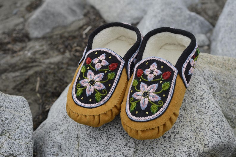 May include: A pair of brown leather moccasins with floral embroidery. The embroidery features pink, white, and green flowers on a black background.