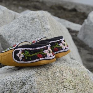 May include: A pair of black moccasins with floral embroidery. The moccasins are made of leather and have a soft, suede-like texture. The embroidery features pink, red, and green flowers and leaves.