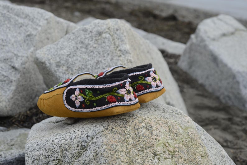 May include: A pair of black moccasins with floral embroidery. The moccasins are made of leather and have a soft, suede-like texture. The embroidery features pink, red, and green flowers and leaves.