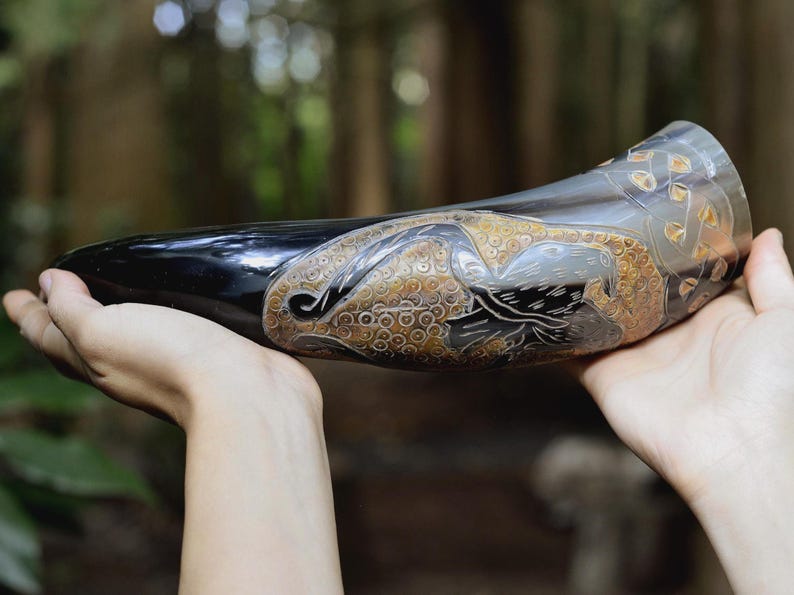 May include: A polished, curved drinking horn featuring intricate, hand-carved details of a bird in flight against a textured, golden-brown background. The horn transitions from a deep, glossy black at the pointed tip to a translucent, light gray at the rim.