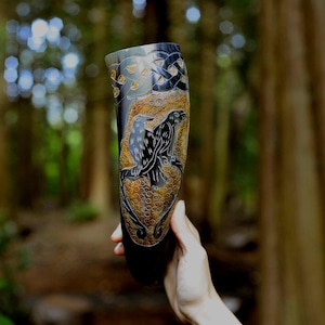 May include: A dark, polished drinking horn featuring intricate carvings of two ravens set against a textured, golden-brown background. The upper portion displays a black Celtic knotwork pattern, while the lower section shows circular motifs.