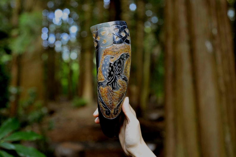 May include: A dark, polished drinking horn featuring intricate carvings of two ravens set against a textured, golden-brown background. The upper portion displays a black Celtic knotwork pattern, while the lower section shows circular motifs.