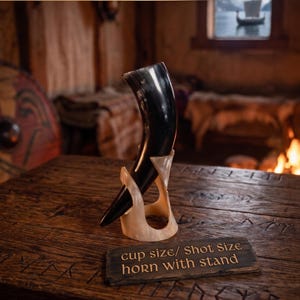 May include: A dark, polished drinking horn with a curved shape, supported by a light-colored, translucent horn stand. A small rectangular plaque in front of the stand features the text: cup size/ Shot Size horn with stand.