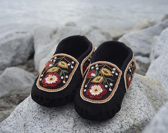 Puede incluir: Un par de mocasines negros con intrincados bordados florales en tonos rojos, amarillos y blancos. Los mocasines están hechos de cuero suave y flexible y tienen un diseño tradicional.