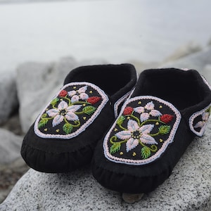 May include: A pair of black moccasins with floral beadwork designs. The flowers are pink, white, and green. The moccasins are on a rocky surface.