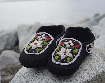 May include: A pair of black moccasins with floral beadwork designs. The flowers are pink, white, and green. The moccasins are on a rocky surface.