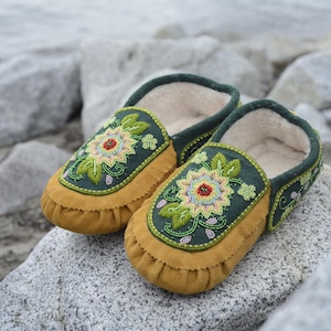 May include: A pair of brown leather moccasins with green and yellow floral embroidery. The moccasins are lined with white fur and have a soft, comfortable feel.