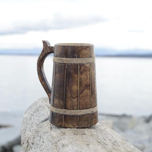May include: A wooden beer stein with a brown finish and a rope handle. The stein is sitting on a piece of driftwood.
