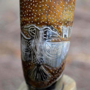 May include: A cylindrical drinking horn featuring a carved owl design with intricate line work and a stippled texture. The surface displays natural brown and tan color variations with deep grooves highlighting the avian figure and geometric patterns.