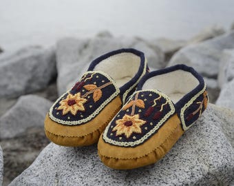 May include: A pair of brown leather moccasins with intricate floral embroidery in shades of yellow, red, and blue. The moccasins have a soft, plush lining and are ideal for keeping your feet warm and cosy.