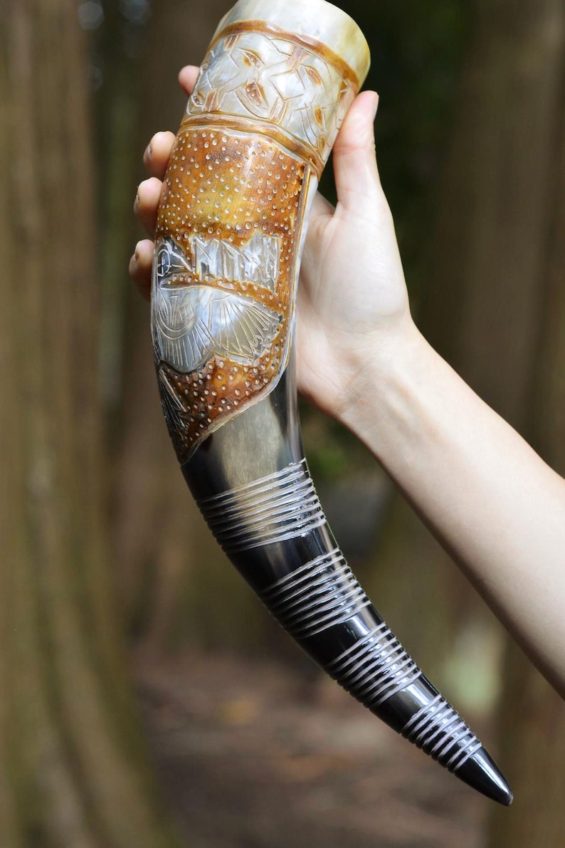May include: A curved drinking horn featuring a polished dark base with horizontal carved rings and a lighter, textured upper section. The upper portion displays intricate etched designs of a bird and geometric patterns in shades of tan and cream.