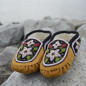 May include: A pair of brown leather moccasins with intricate floral beadwork in shades of pink, white, and green. The moccasins have black trim and are set against a backdrop of gray rocks.